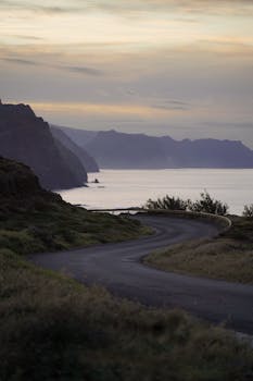 A winding road by the coast with mountains and sea under a sunset sky, perfect for travel inspiration.