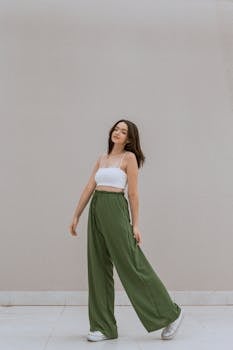 Trendy young woman posing against a grey studio background in green trousers and white crop top.