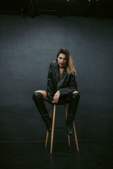 Fashion portrait of a woman in black clothes sitting on a stool against a dark background.