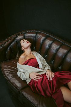 Woman in a red silk dress lying elegantly on a leather sofa with eyes closed, exuding fashion and sensuality.