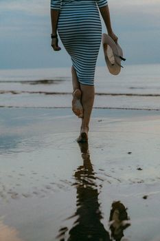 Back view of woman in striped dress walking barefoot on beach, holding hat at dusk.