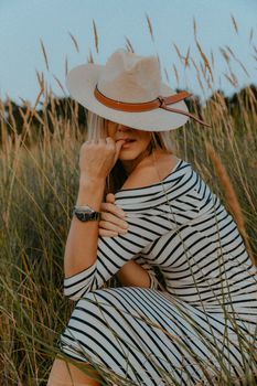 Blonde woman in a hat posing in a field wearing a striped dress. Fashion and nature merge.