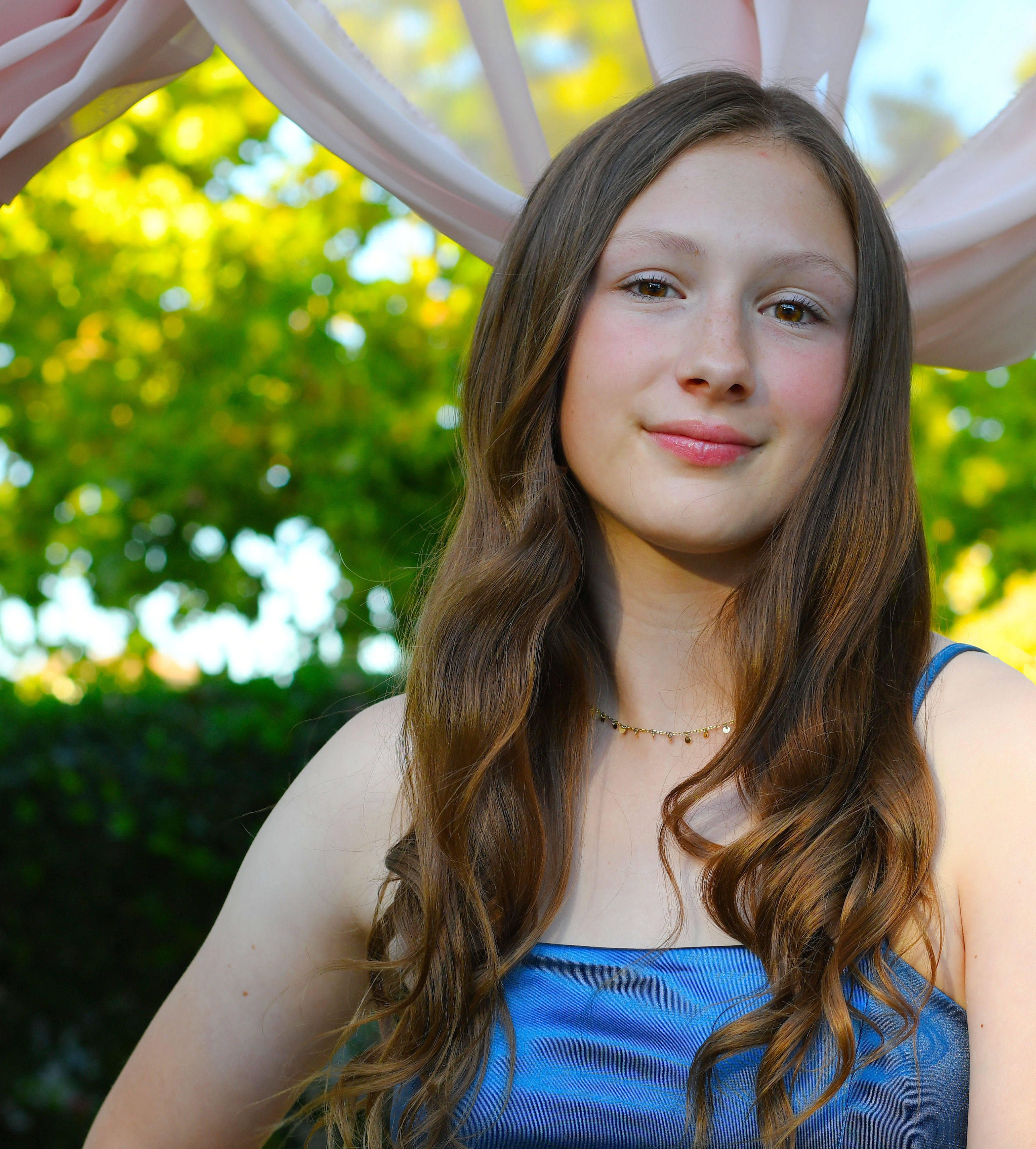 A teenage girl smiling joyfully in a blue dress outdoors, in a sunlit park setting.