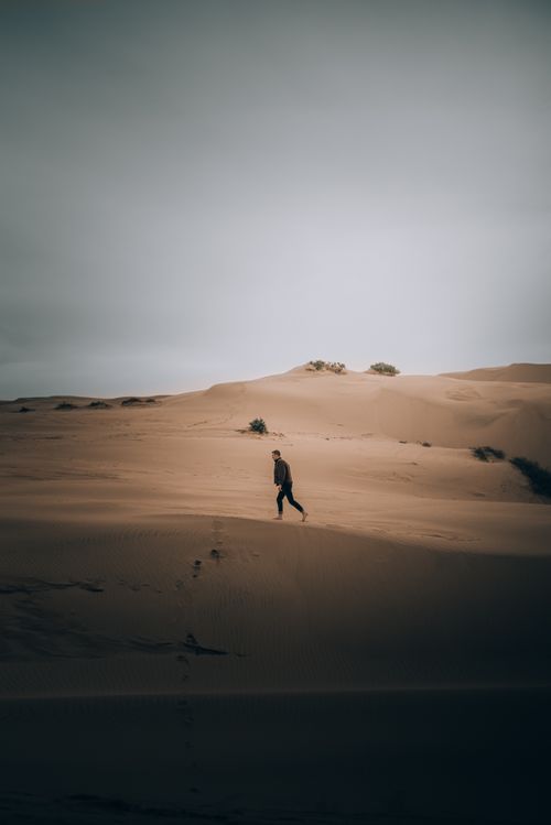 Free A solitary person walking through expansive desert sand dunes under a cloudy sky. Stock Photo
