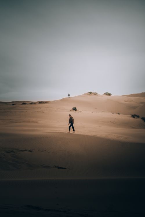 Free Silhouetted man walking across vast desert dunes under a dramatic sky at sunset. Stock Photo