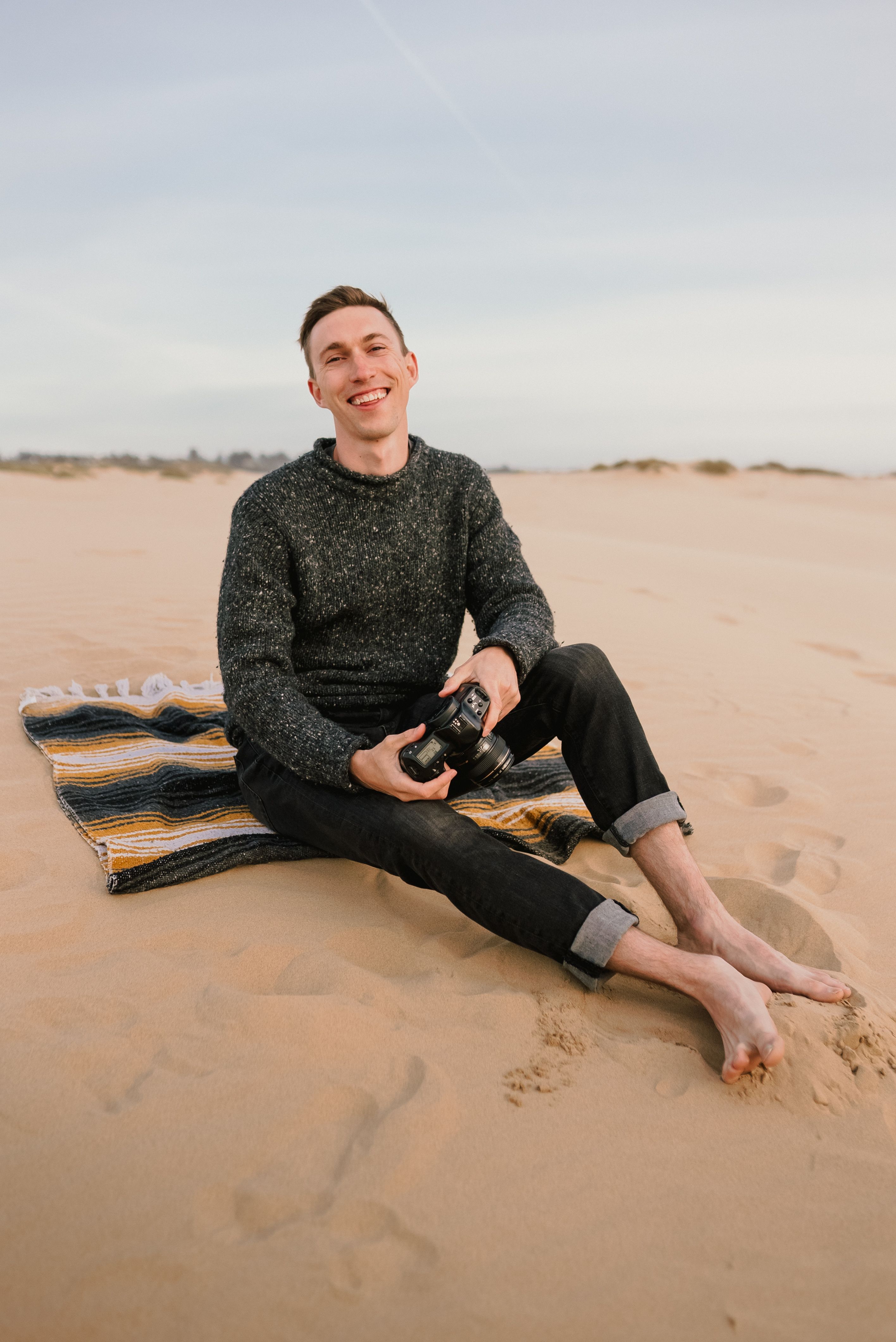 A relaxed man sits barefoot on a beach holding a camera, enjoying a sunny day.