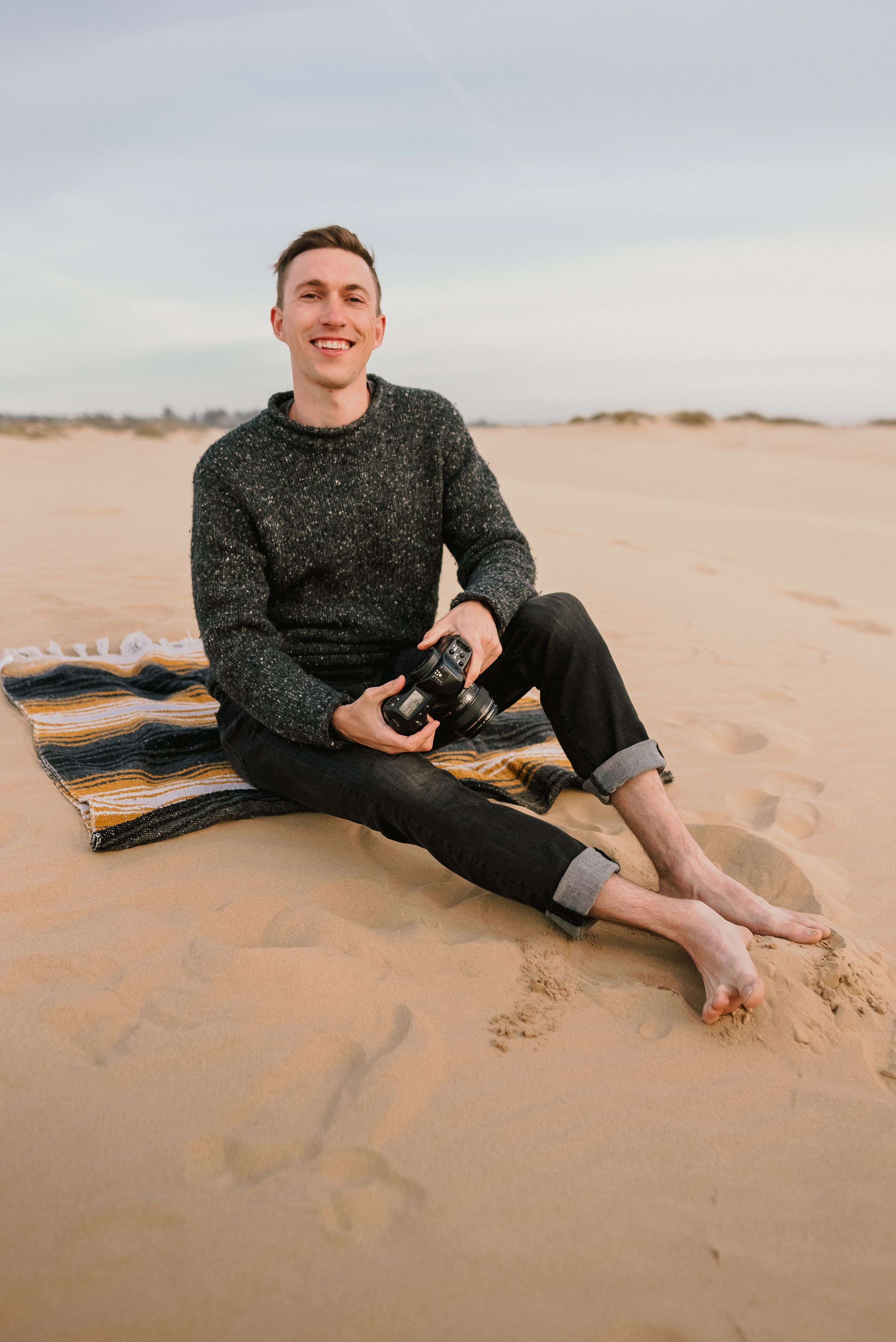 A smiling man sitting barefoot on a sandy beach, holding a camera.