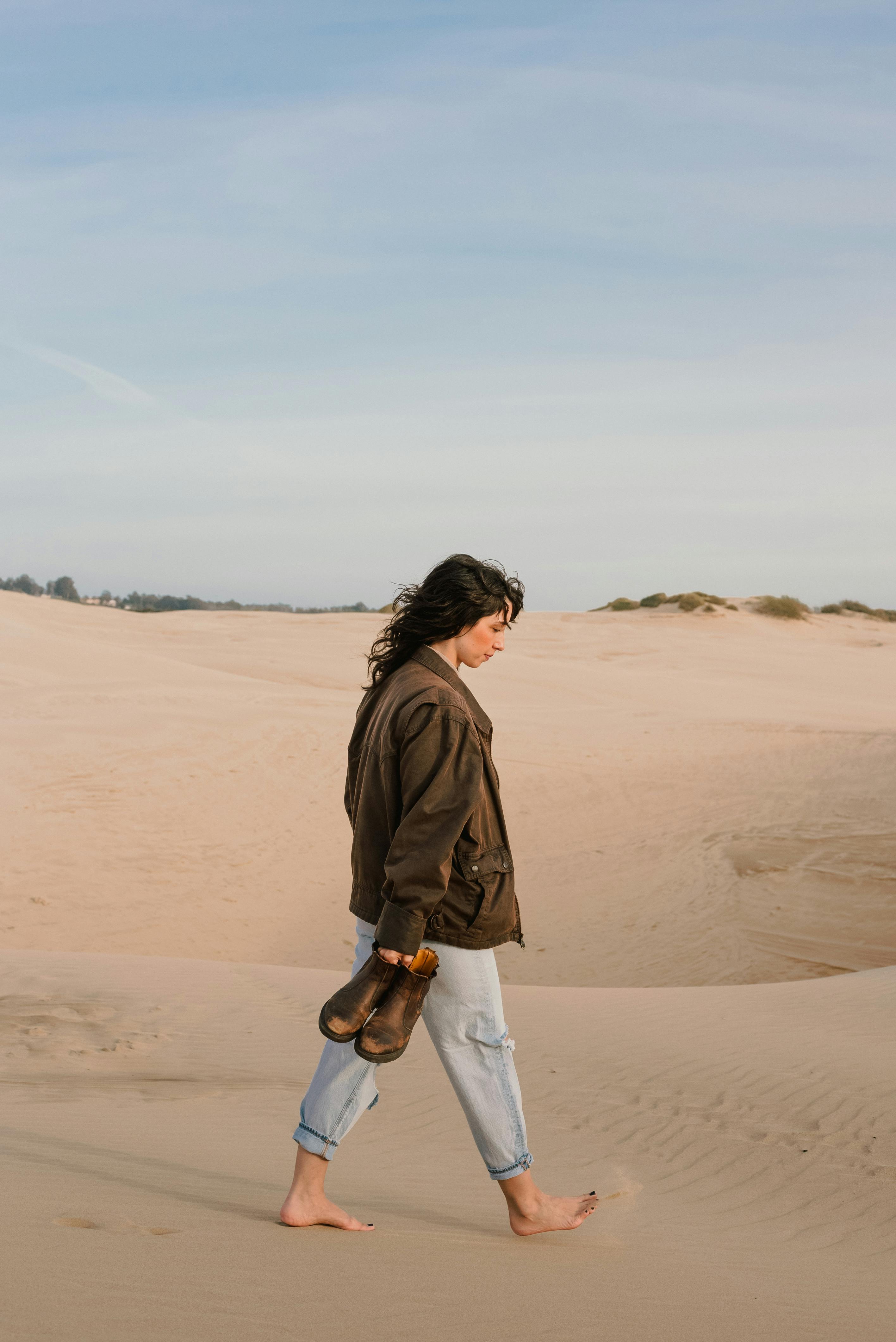 A woman walks barefoot on sandy dunes under a clear blue sky, carrying shoes.