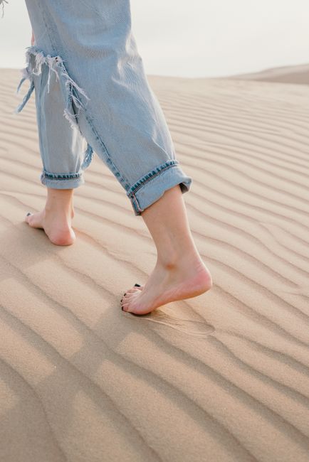 A woman walks barefoot on sandy beach dunes, showcasing torn jeans and summer vibes.