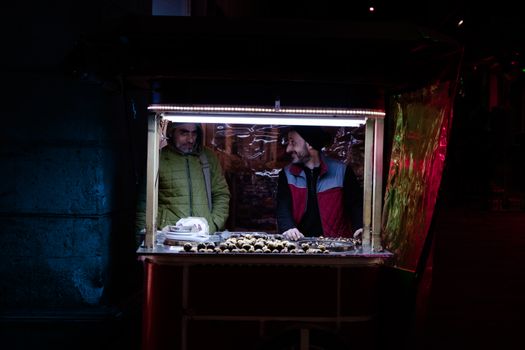 Street vendors at a night market stall, illuminated by neon lights, offering snacks.