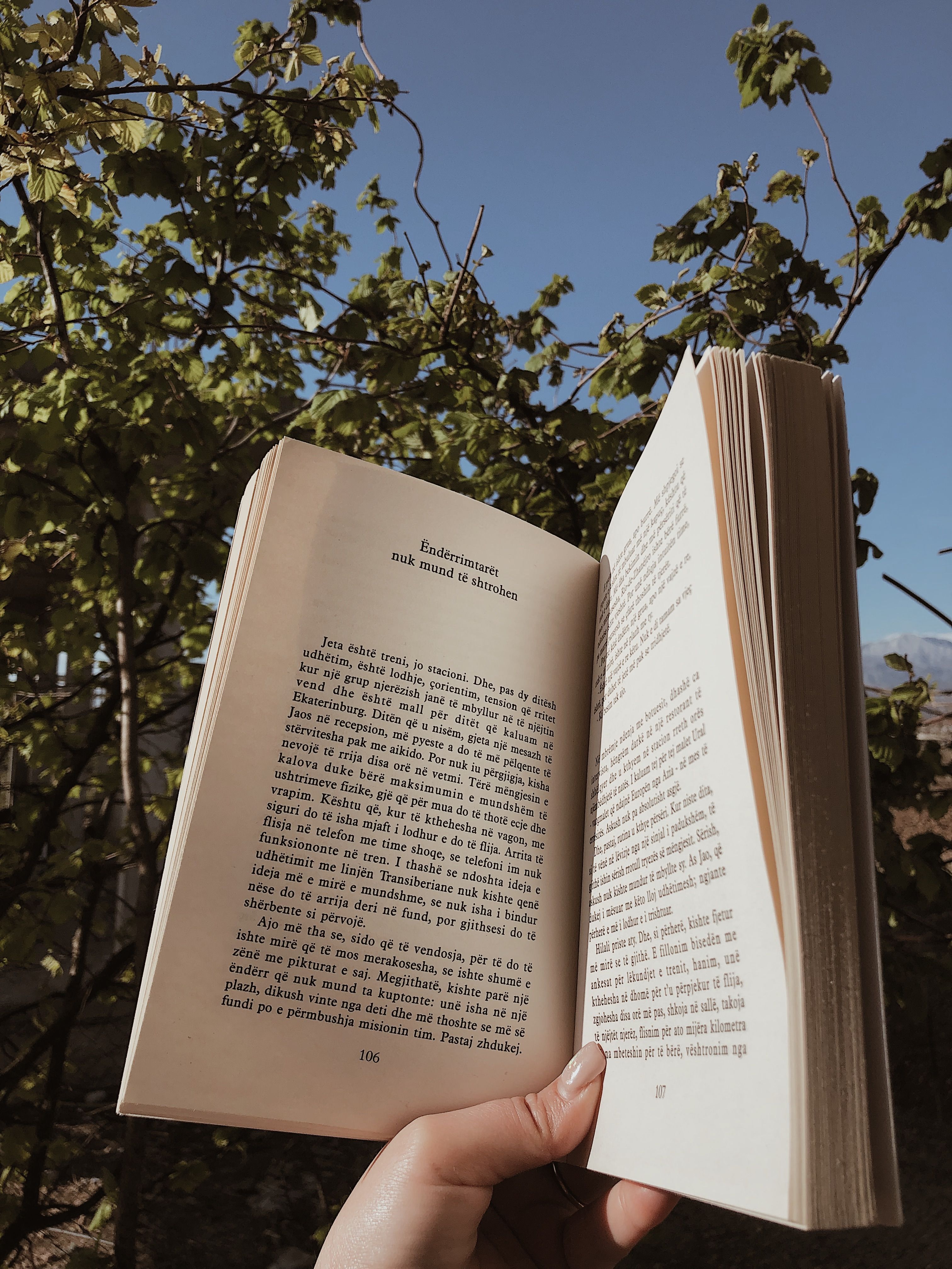 Hand holding an open book against a backdrop of greenery and clear sky.