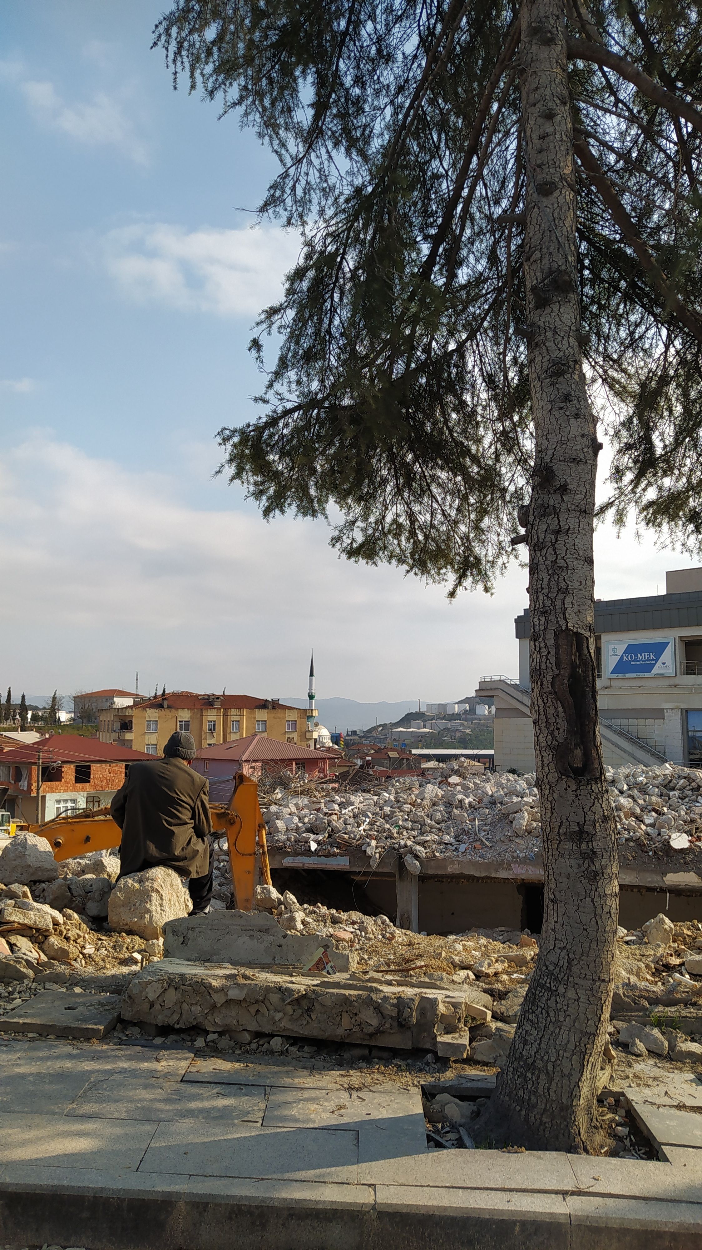 A man sits by an excavator amid urban demolition and scattered debris.