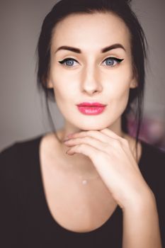Thoughtful young female in black shirt with tied dark hair and grey eyes wearing eyeliner makeup holding hand under chin