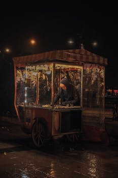 A man serves food at a vibrant street food cart at night, highlighted by city streetlights.