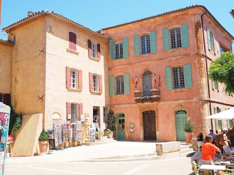 A sunny day in a picturesque French village square with colorful buildings and lively atmosphere.