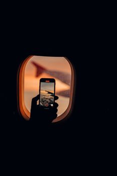 Silhouette of a person filming sunset through an airplane window using a smartphone.