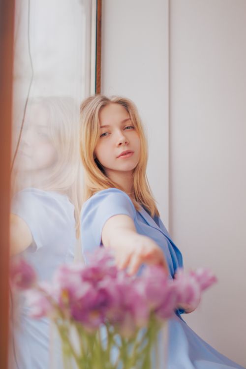 Free Portrait of a young woman in blue sitting by a window with flowers, capturing a serene and thoughtful moment. Stock Photo