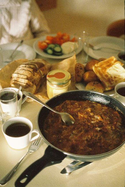 A cozy brunch setting featuring a skillet dish, bread, coffee, and fresh vegetables on a sunny table.