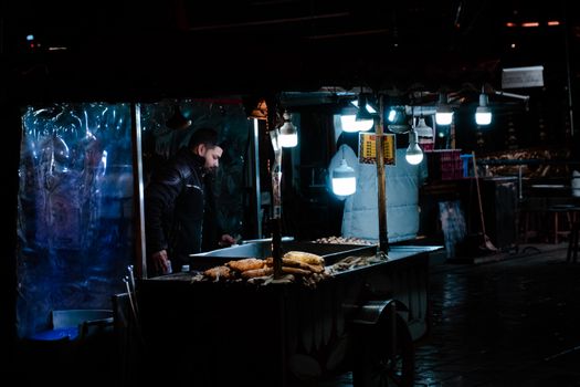 A street food vendor serves grilled corn at night, illuminated by hanging bulbs.
