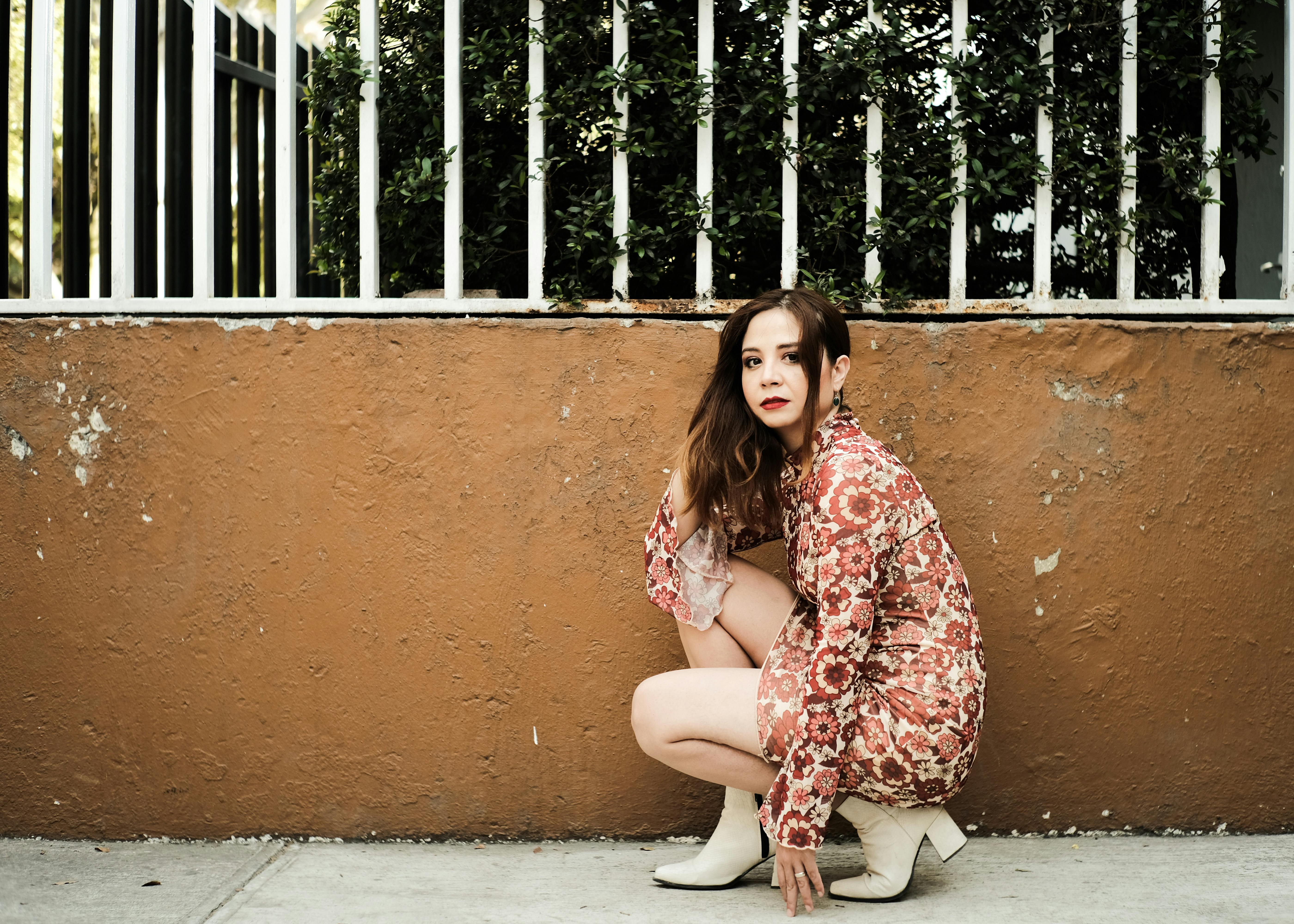 A stylish woman crouching by a wall in an urban setting, wearing a patterned dress and boots.
