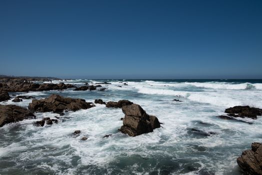 Stunning California coast with crashing waves and rugged rocks under clear blue skies.