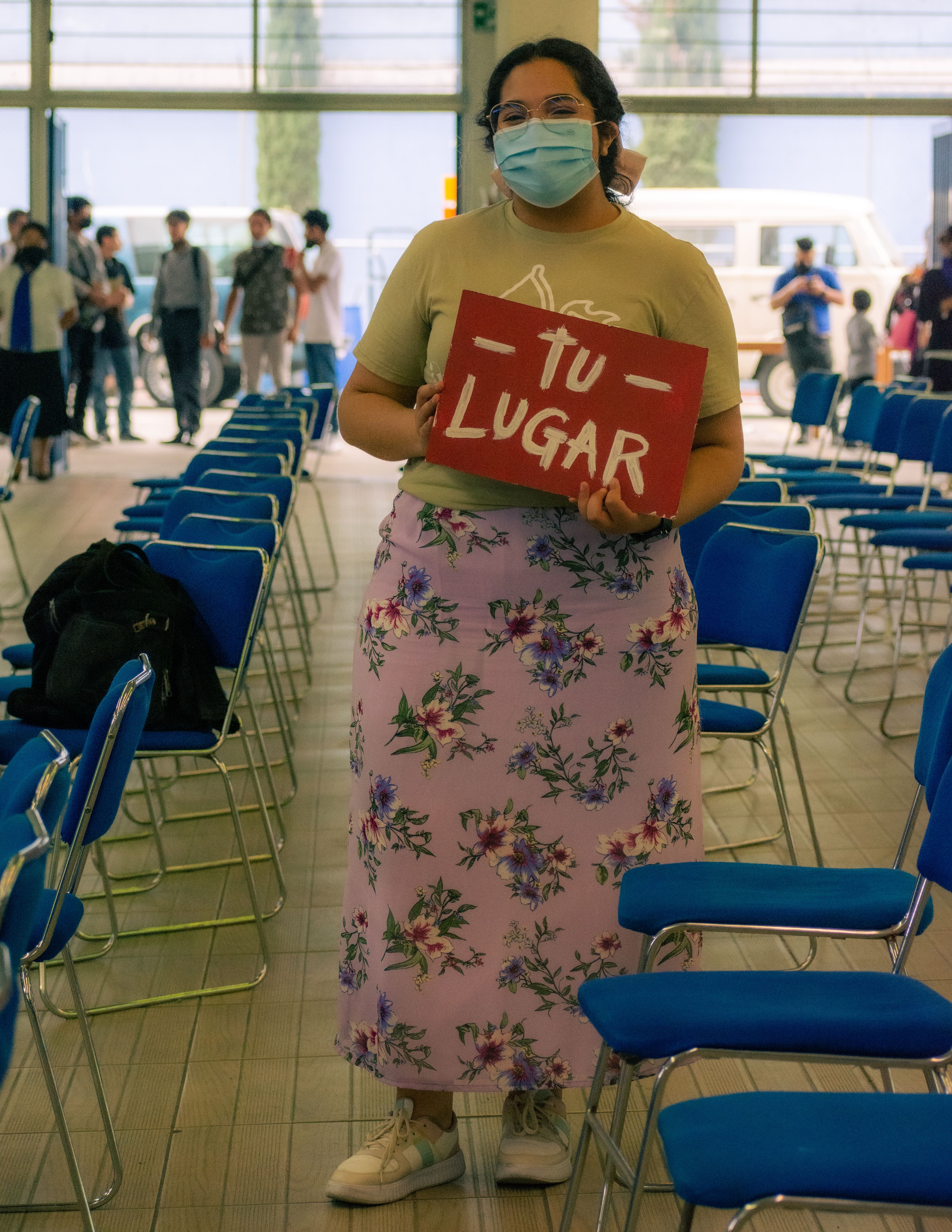 A woman holds a 'Tu Lugar' sign at an indoor event with rows of chairs.