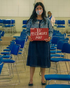 A woman in a mask holds a sign reading 'Tu Perteneces Aqui' among blue chairs indoors.