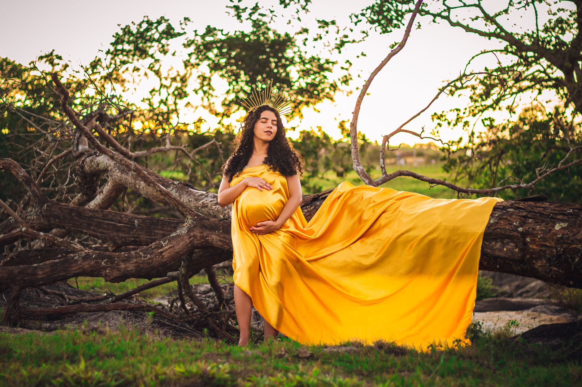 A serene image of a pregnant woman in a yellow gown posing outdoors by a fallen tree.
