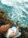一名男子穿着休闲装，坐在岩石悬崖上，平静地凝视着下方的海浪