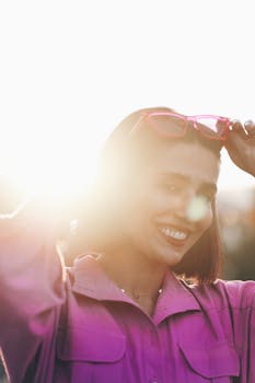 A cheerful young woman in pink adjusts her sunglasses as sunlight creates a warm, glowing effect.