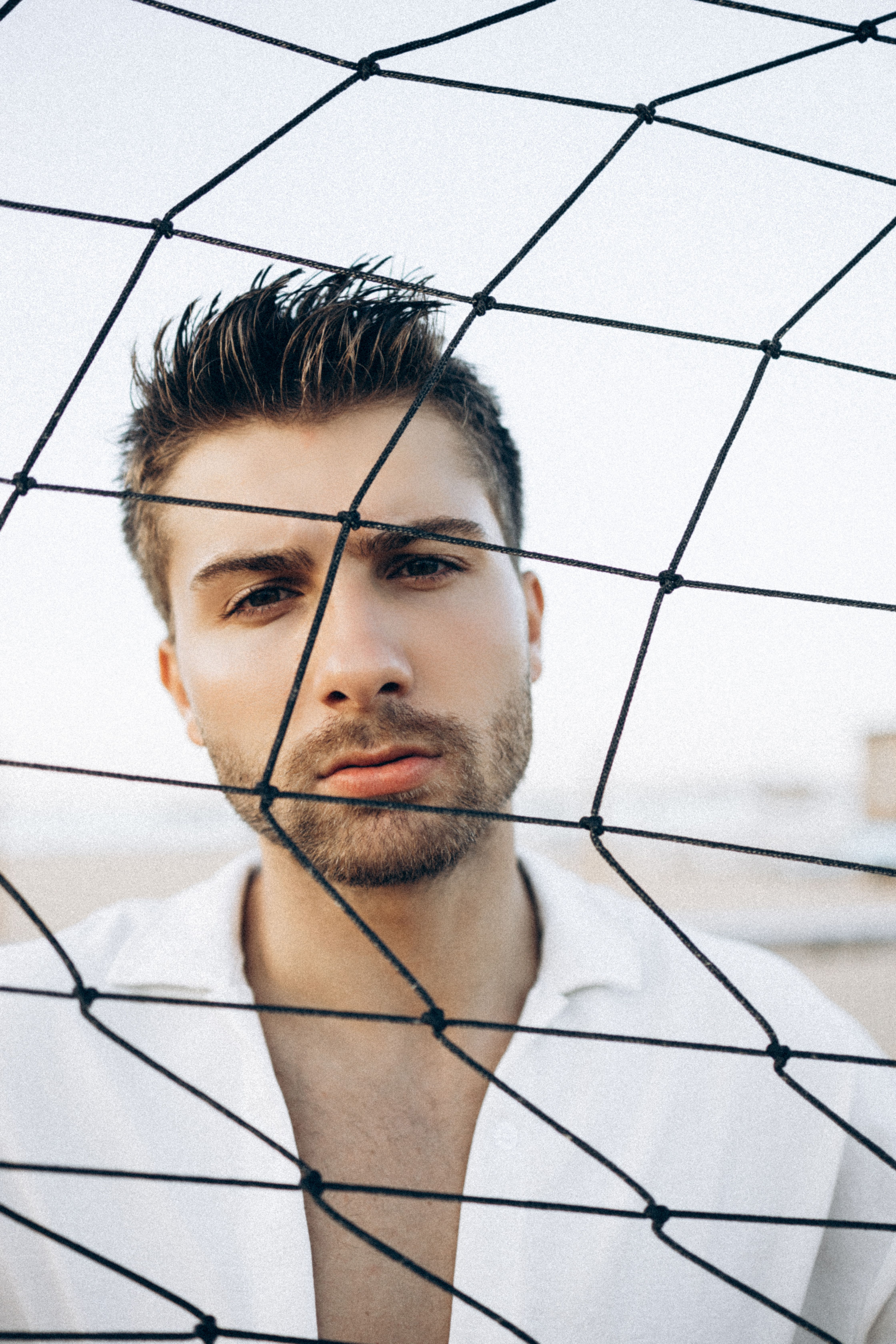 Portrait of a young man with stubble behind a net, outdoors. Modern style and intense gaze.