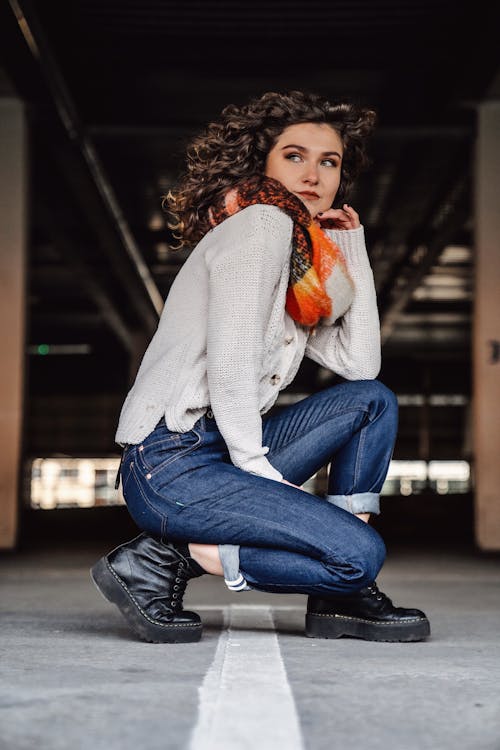 Photo of a Young Woman in Jeans and a Sweater Crouching in a Parking Lot