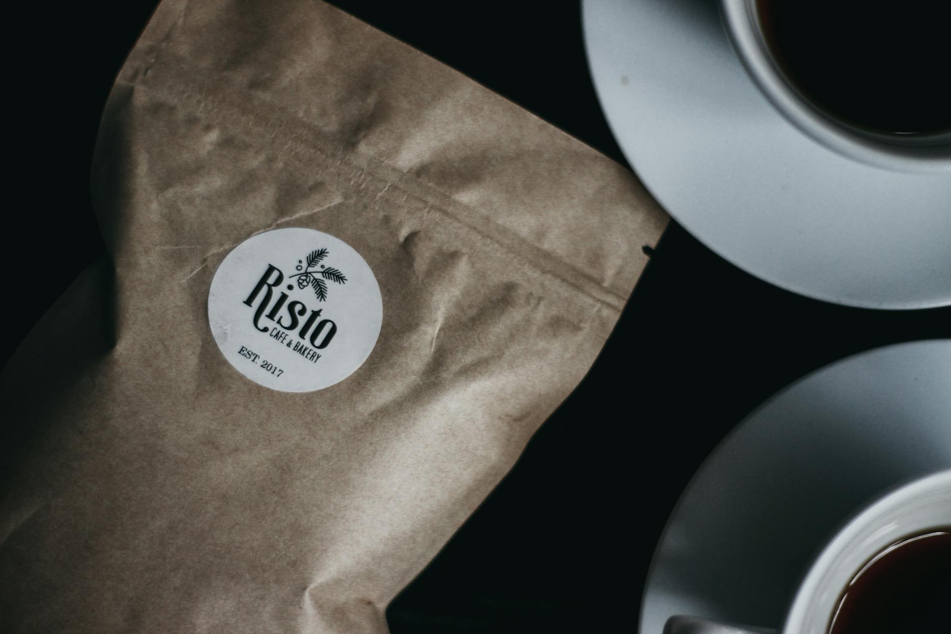 Close-up of a coffee bag labeled 'Risto' next to two coffee cups on a dark surface.