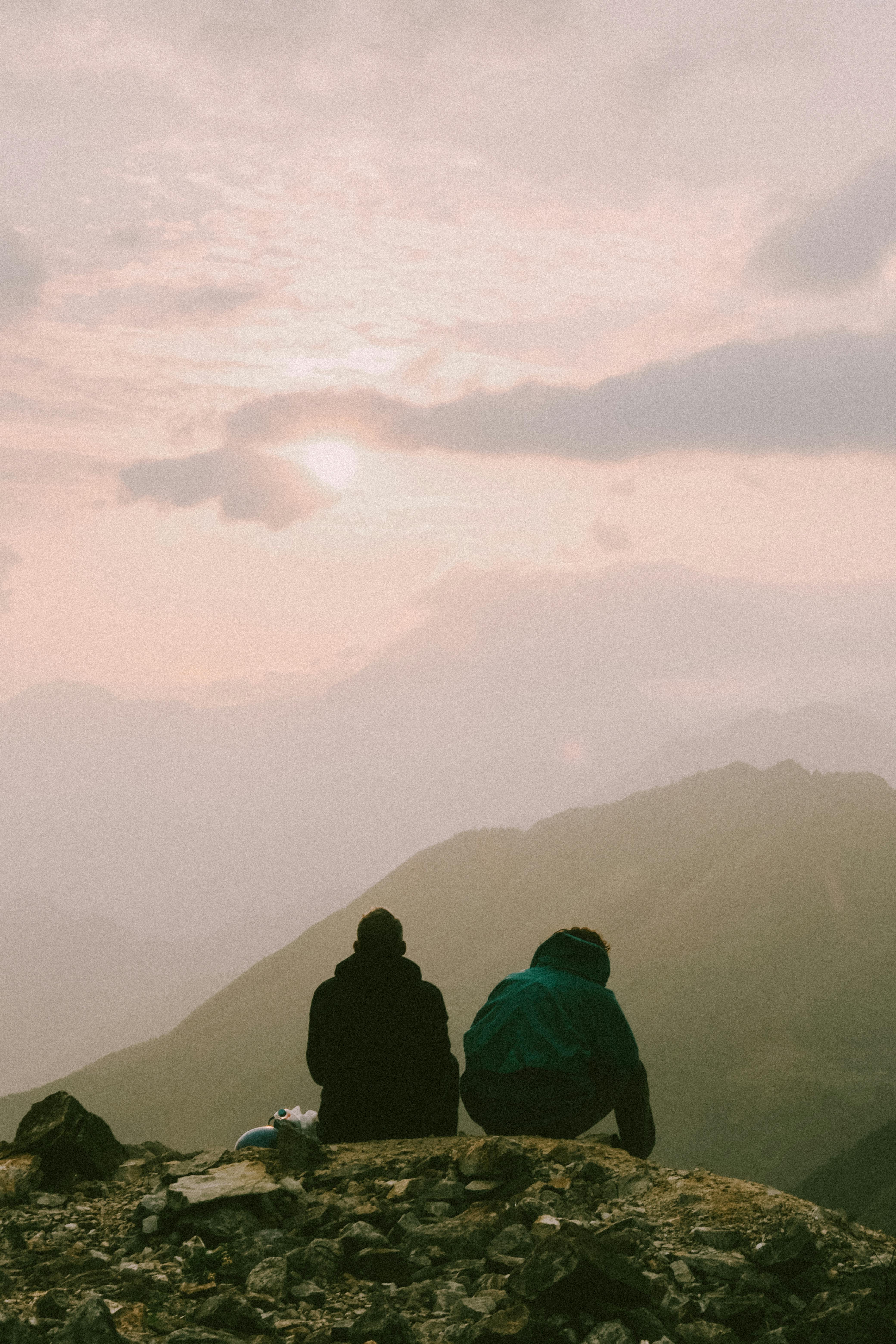 Two people seated on a rocky hill, admiring a serene mountain landscape during sunset.