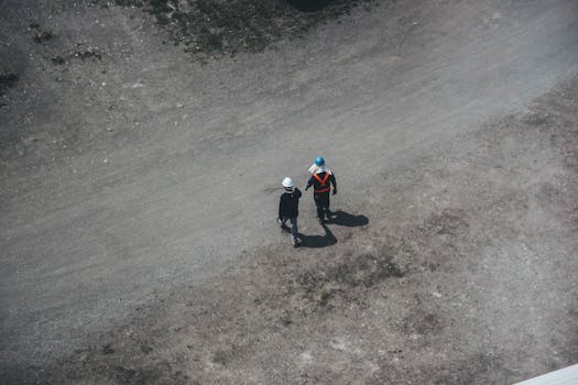 Two construction workers walking on a gravel road with safety gear and helmets, seen from above.
