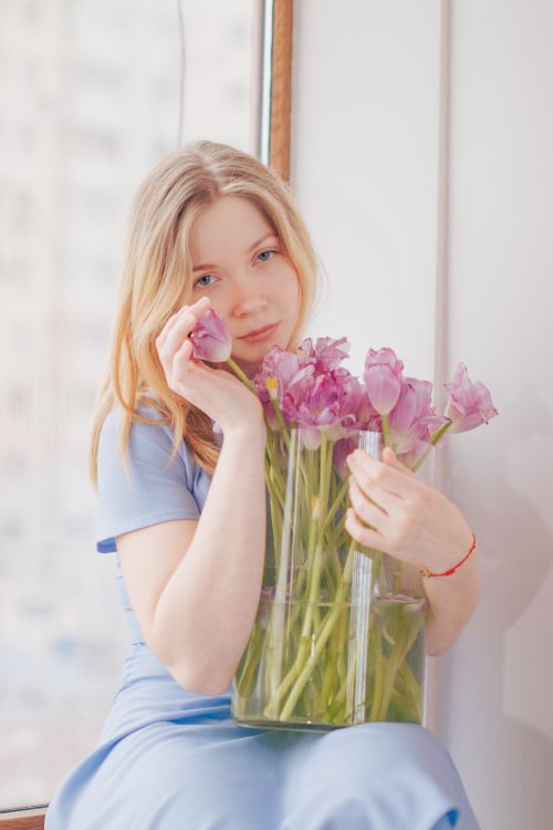 Free Blonde woman in blue dress holding pink tulips, posing by the window in an indoor setting. Stock Photo