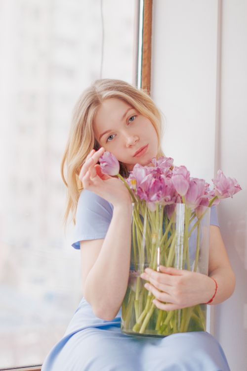 Free A serene portrait of a woman holding pink tulips in a vase by a window indoors. Stock Photo