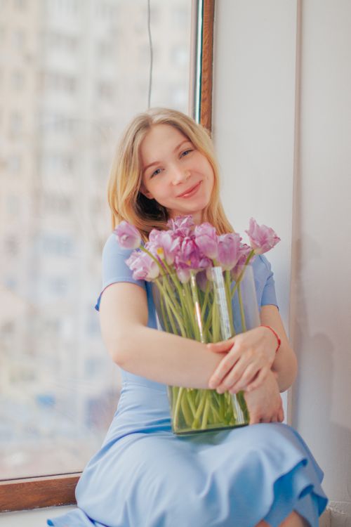 Free Smiling woman in a blue dress holding a vase of pink flowers by a window. Stock Photo