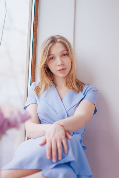Free A serene portrait of a woman with blonde hair sitting by a window, wearing a blue dress. Stock Photo