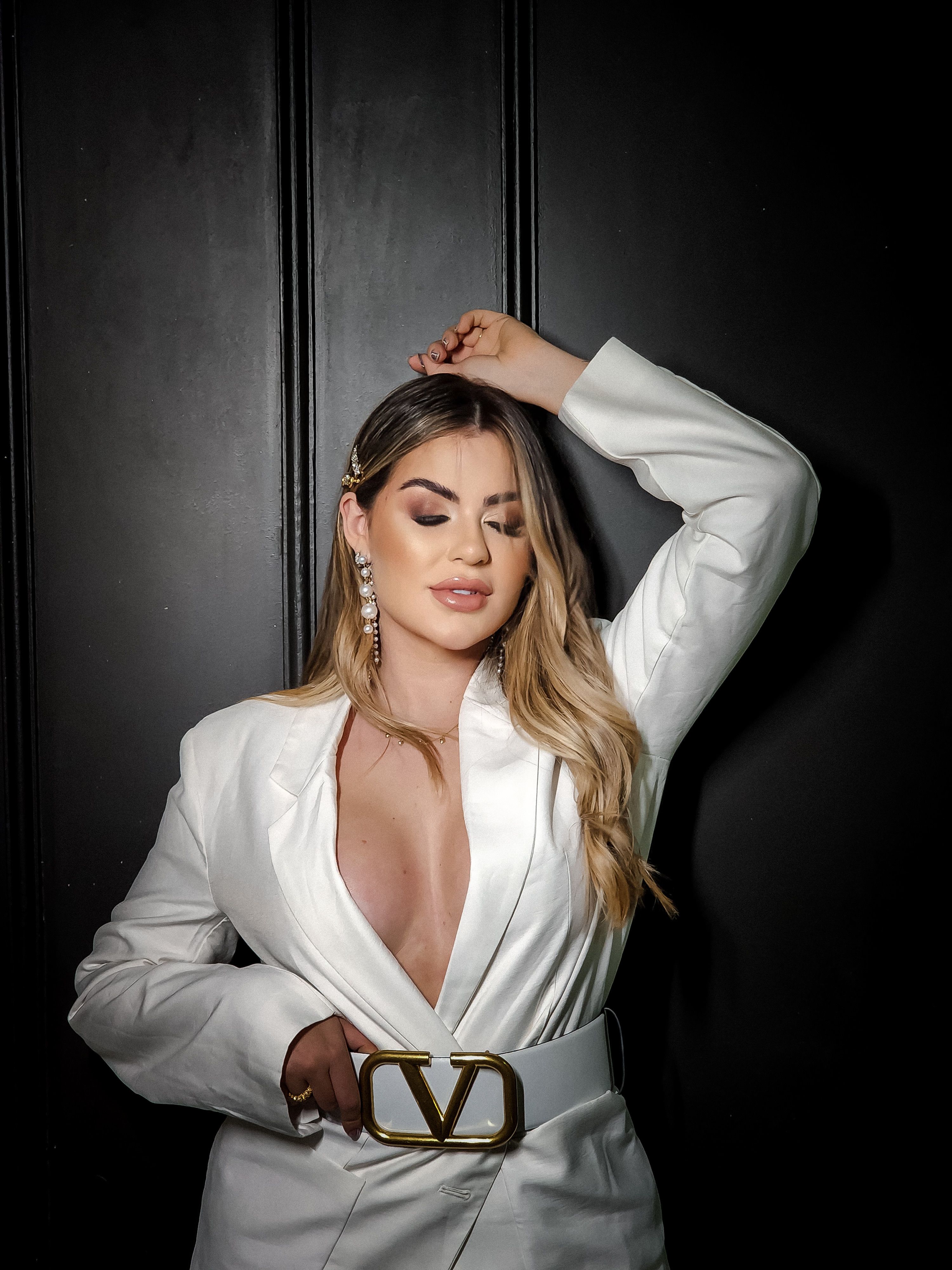 Linda mulher com jaqueta branca posando elegantemente com os olhos fechados contra um fundo escuro.