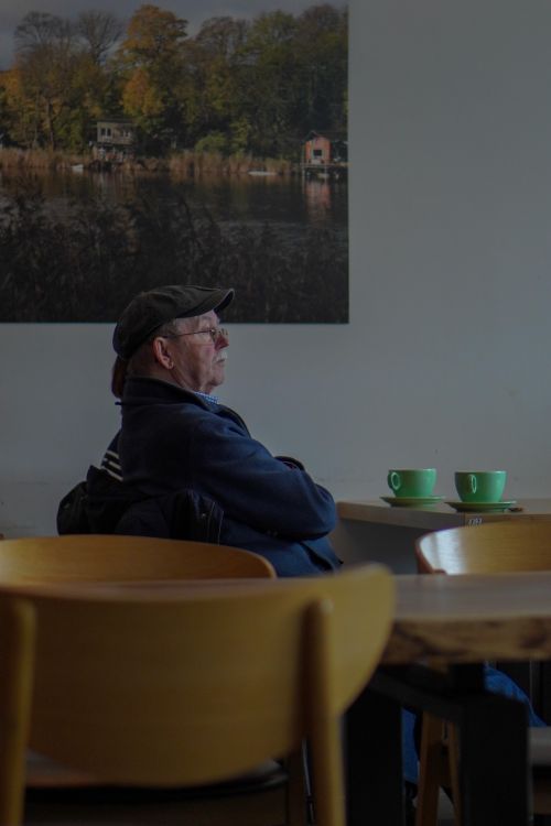 Free An elderly man wearing a cap sits alone in a café, observing his surroundings. Stock Photo