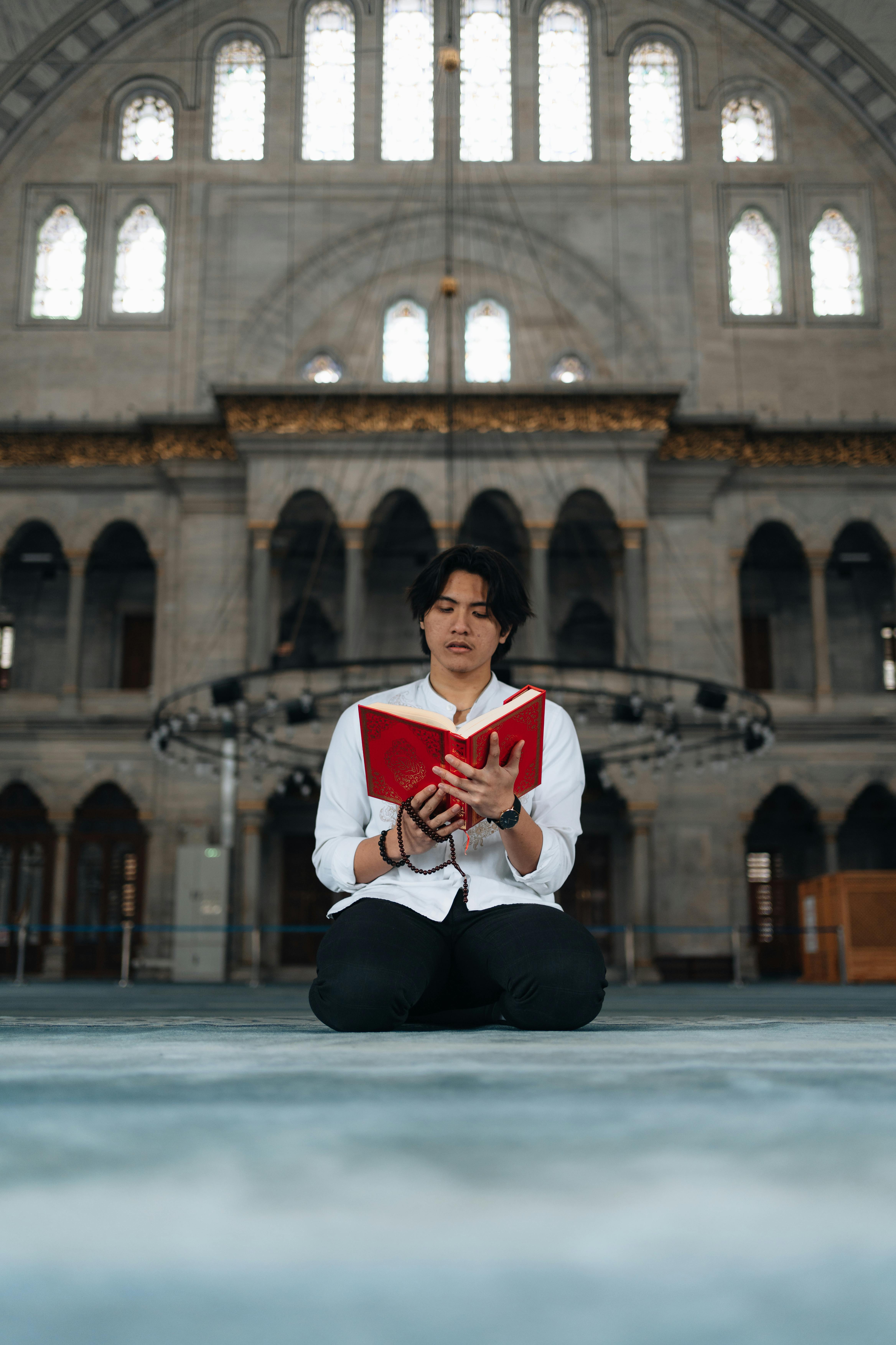 A man in a mosque kneeling and holding a red book, symbolizing faith and spirituality.