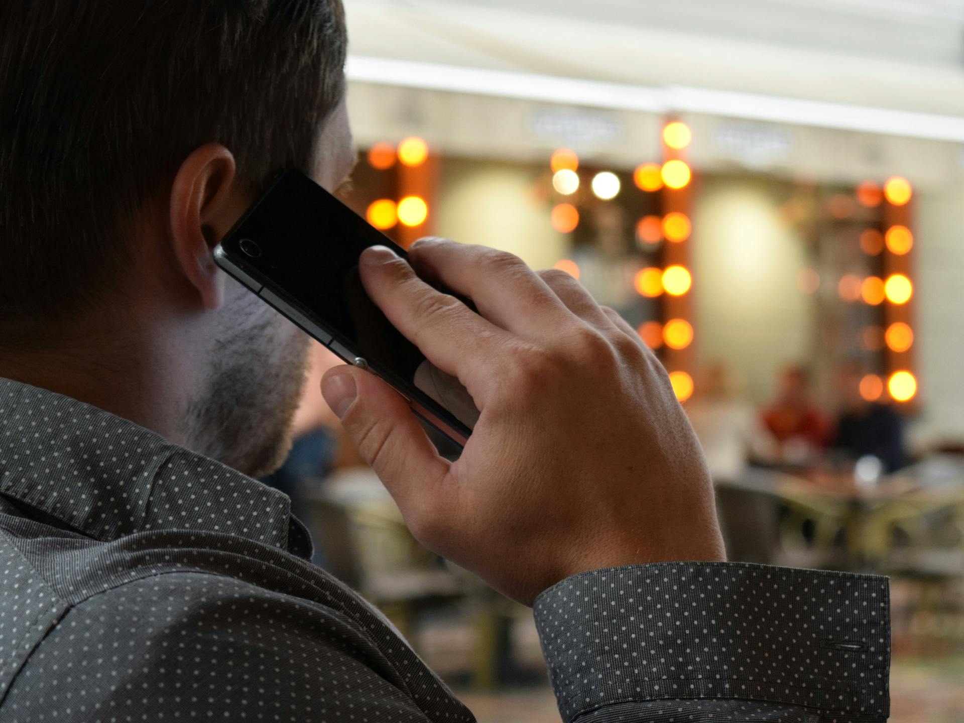 Close-up of a businessman in a dotted shirt making a call in a lively indoor setting with warm lights.