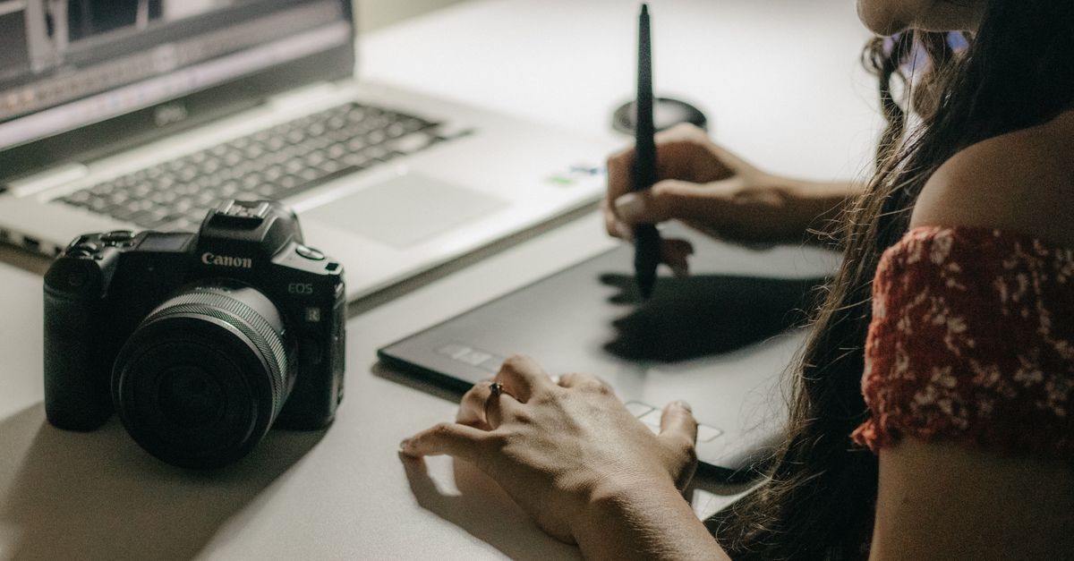 A woman edits photos on a laptop using a tablet and stylus, with a camera beside her.
