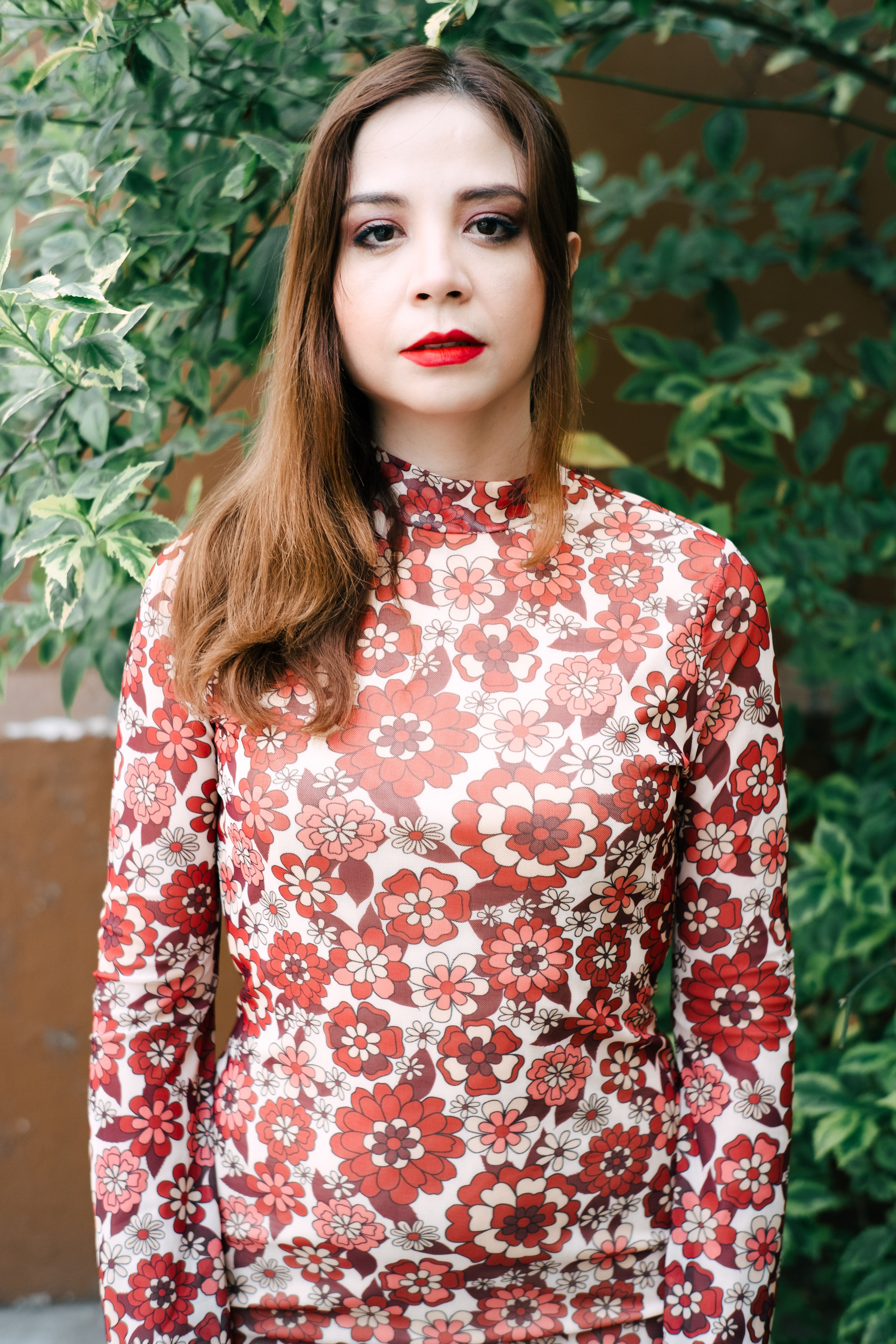 Portrait of a woman in a floral dress with red lips, standing outdoors amidst greenery.