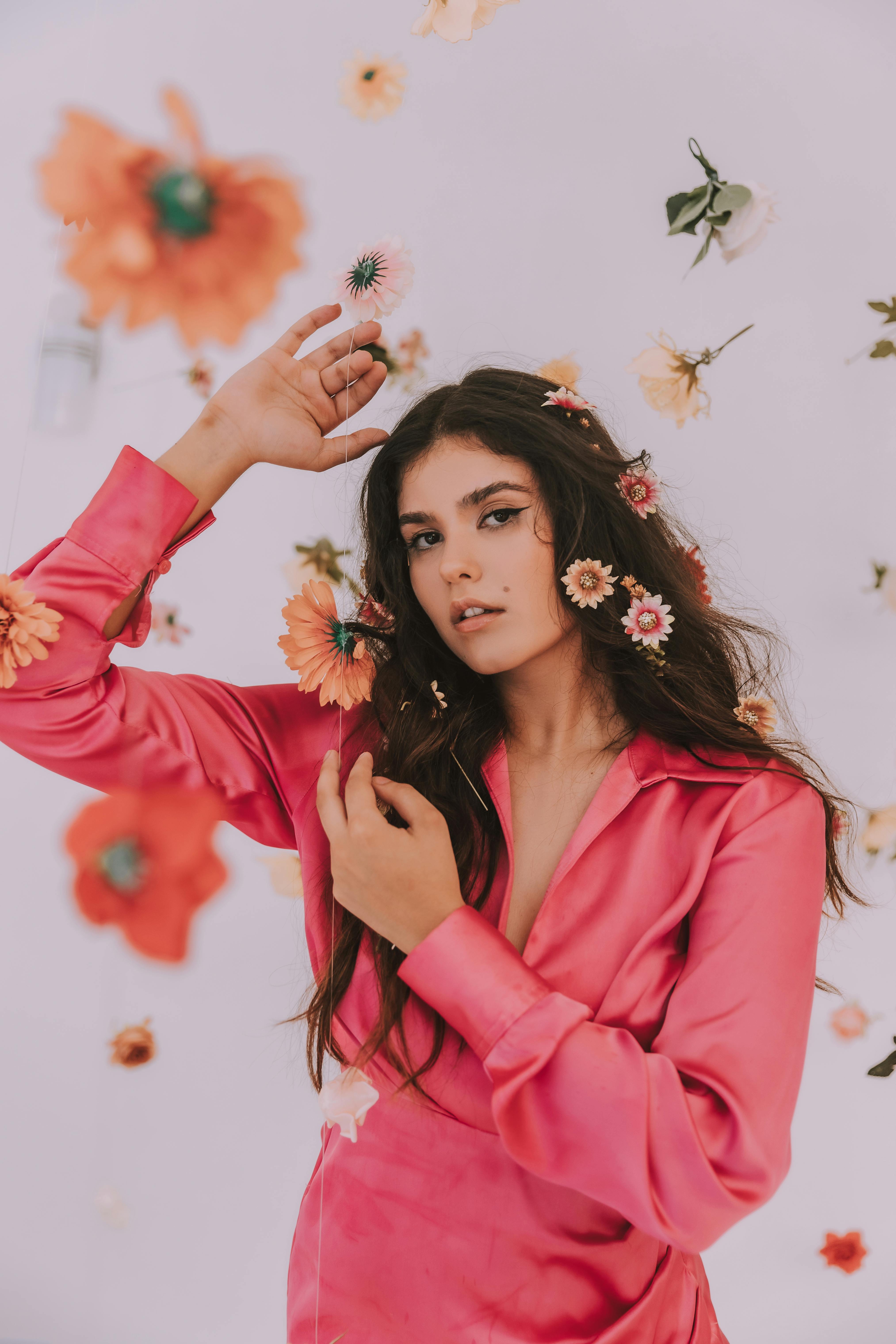 A stylish portrait of a young woman in a pink top surrounded by colorful wildflowers in a studio setting.