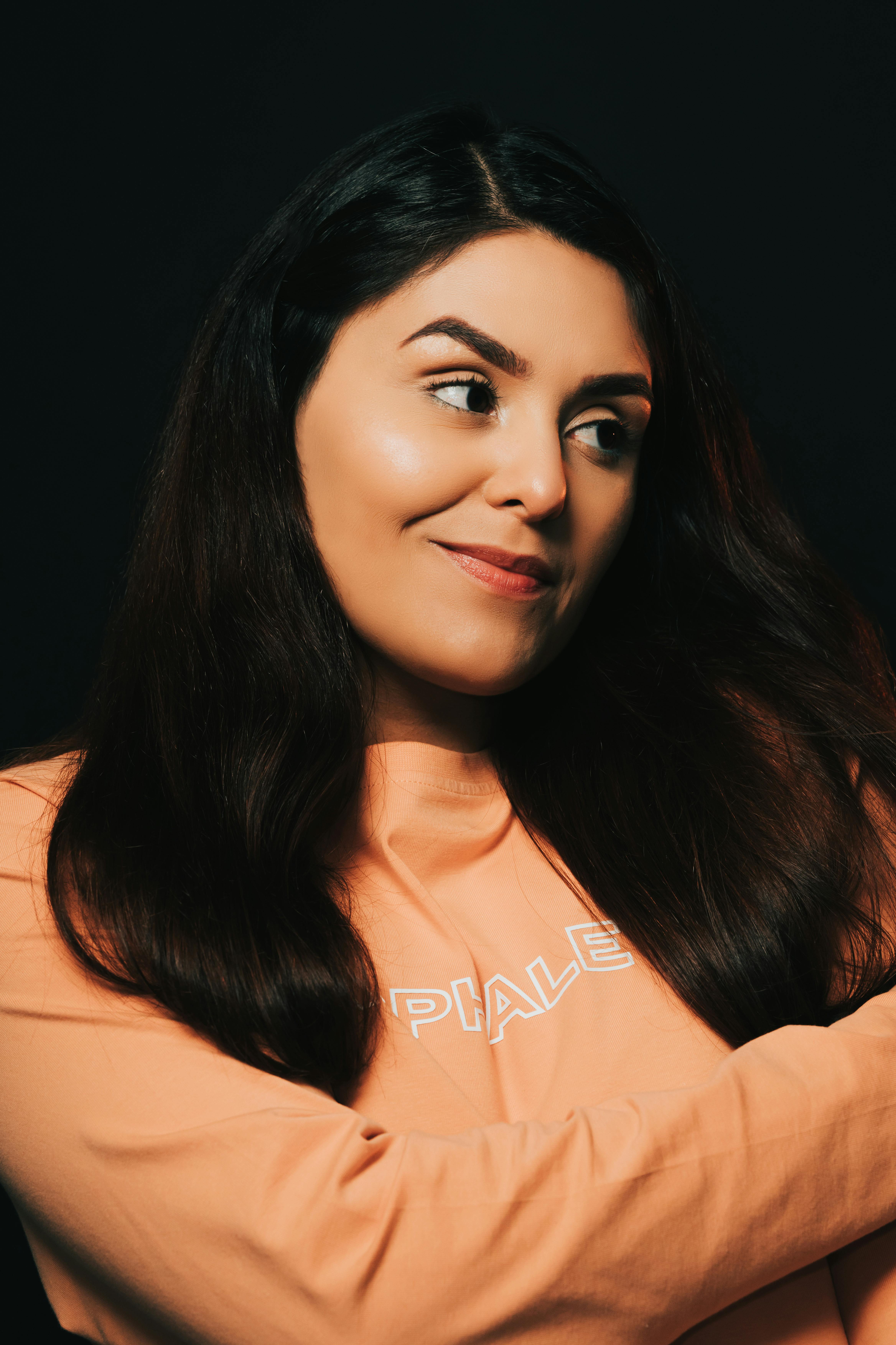 Studio portrait of a woman in an orange sweatshirt with long hair on a dark background.