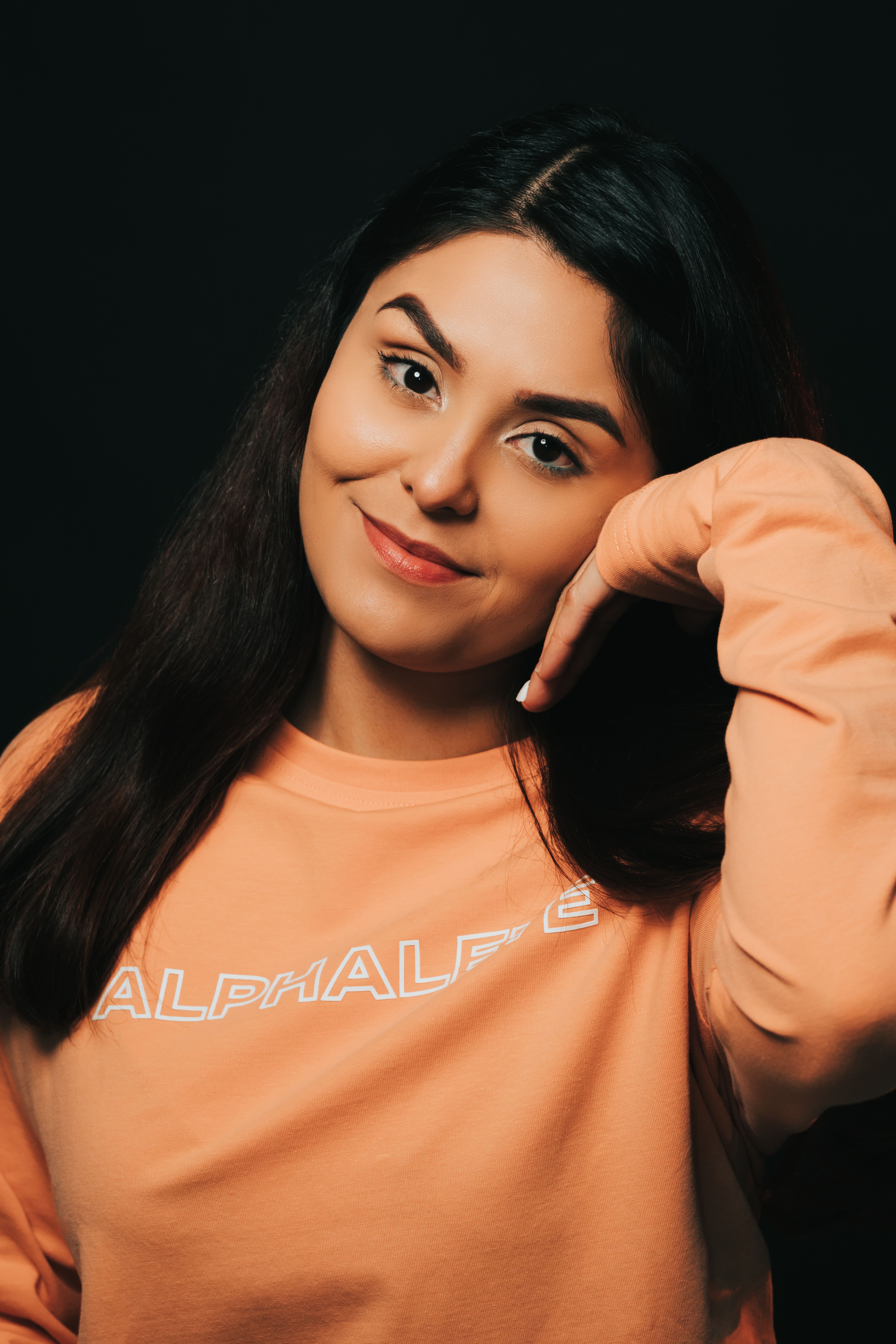 A young woman in a peach sweater posing with a smile against a black background.