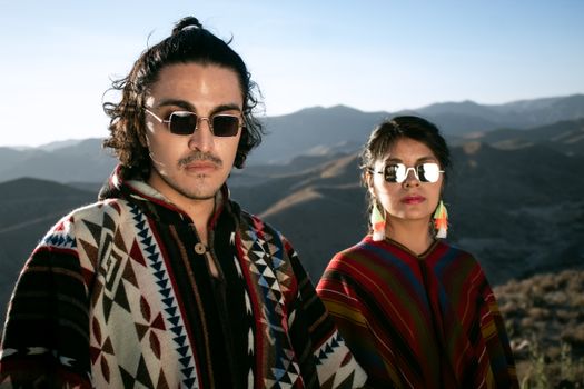 A fashionable couple in colorful ponchos poses with a scenic mountain backdrop, showcasing travel and adventure.