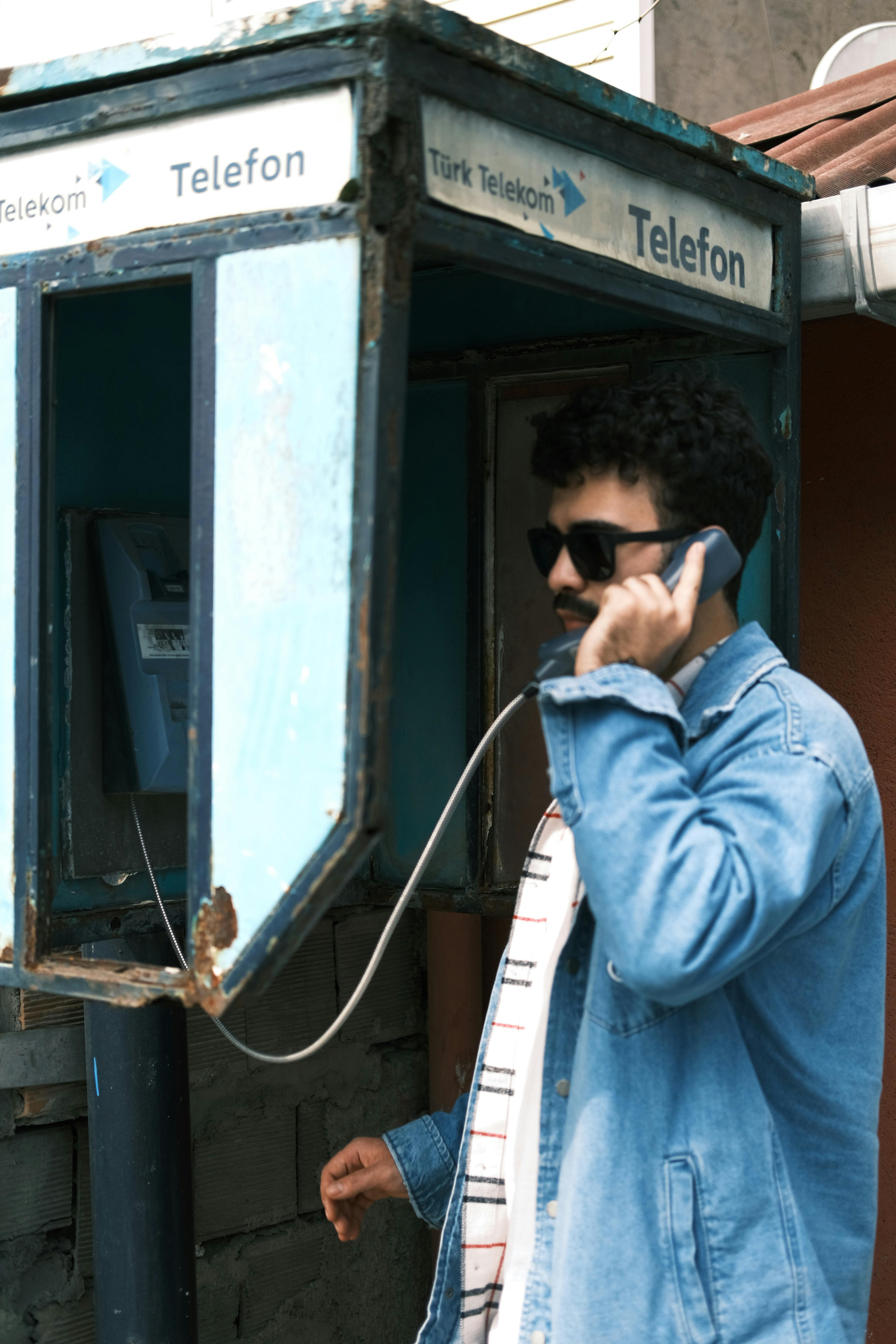 Young man with sunglasses in blue denim interacting with a vintage telephone booth in the city.